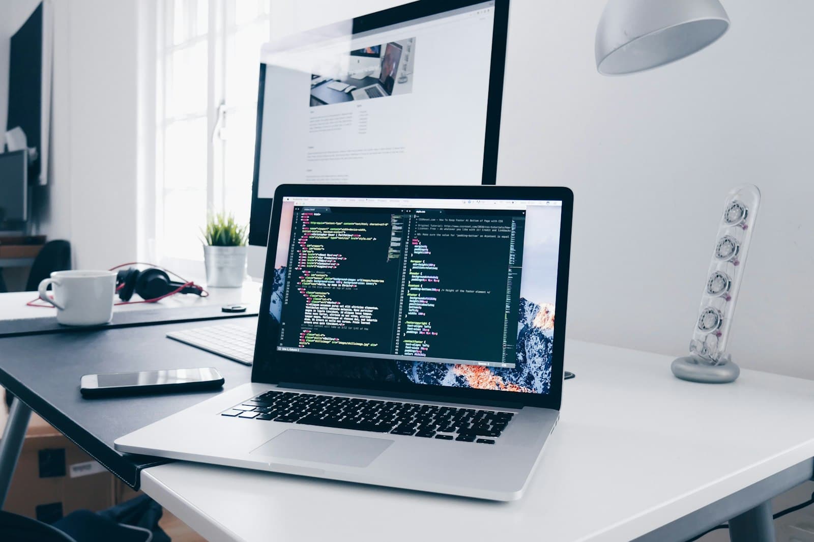 Meja kerja minimalis dengan laptop menampilkan kode editor dan monitor eksternal menampilkan pratinjau halaman website, ditemani secangkir kopi dan tanaman kecil.